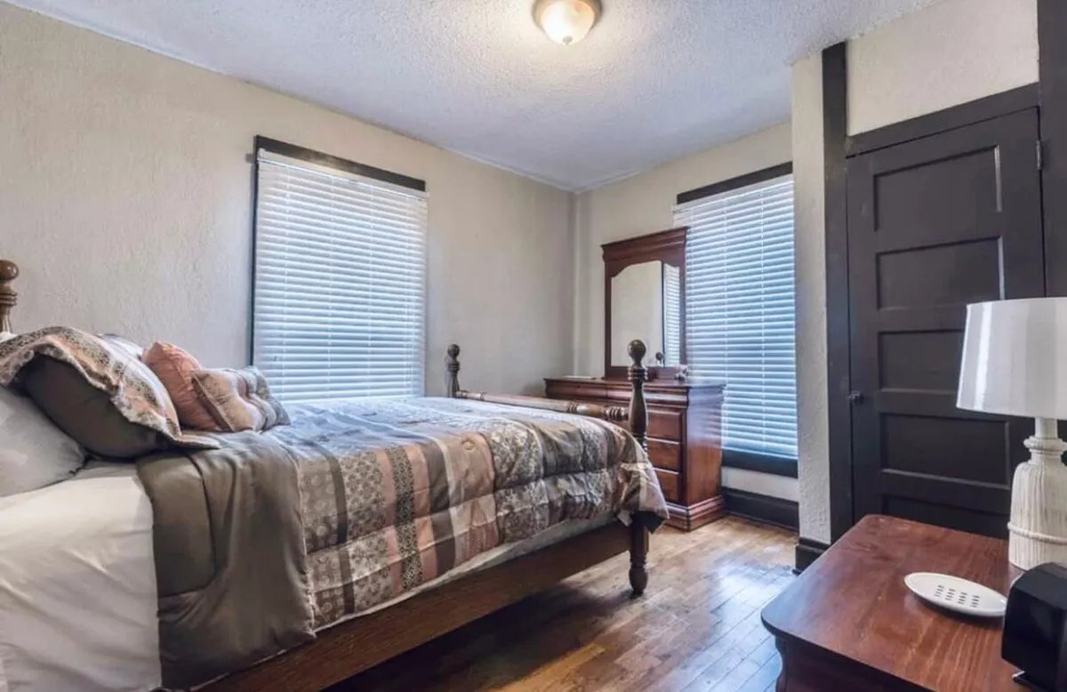 A comfortable bedroom with a stylish patterned quilt on the bed, a wooden dresser, a window with blinds allowing morning light, and a nightstand with a lamp. The space exudes warmth and tranquility, ideal for unwinding after a busy day.