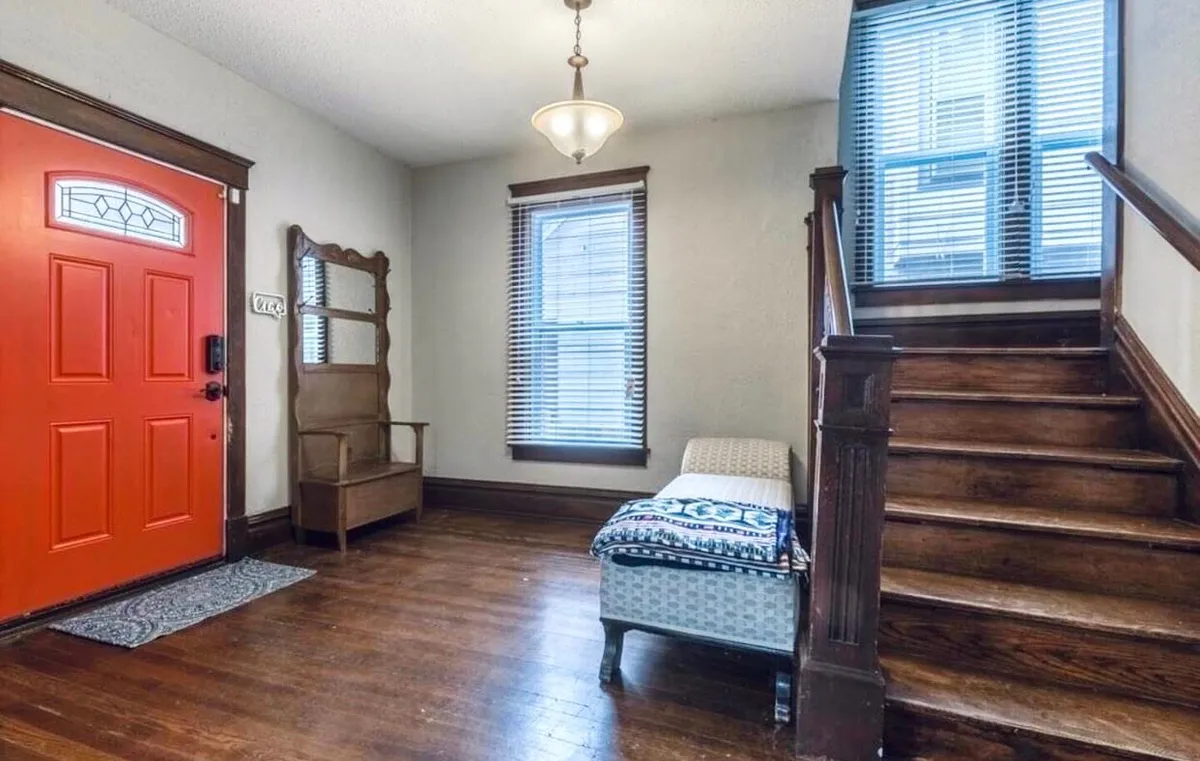 A bright interior entryway featuring a vibrant orange front door, wooden staircase, mirrored coat rack, and a textured seating area. Natural light fills the space, making it an inviting and comfortable spot, perfect for reading by the window or enjoying a morning coffee.