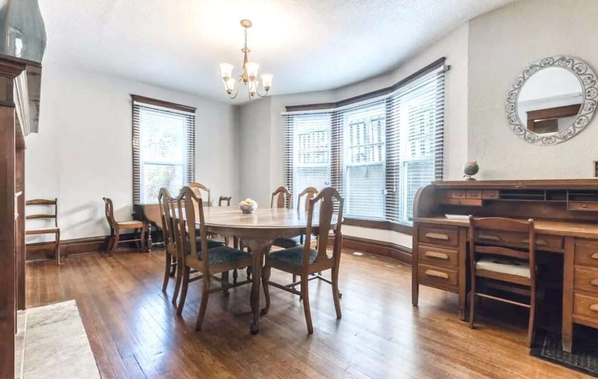 A spacious dining area with a large table set beneath a chandelier, flanked by bay windows that flood the room with warm natural light. There's also a writing desk in the corner, adding versatility to the space. The setting is inviting, perfect for family dinners or quiet moments.