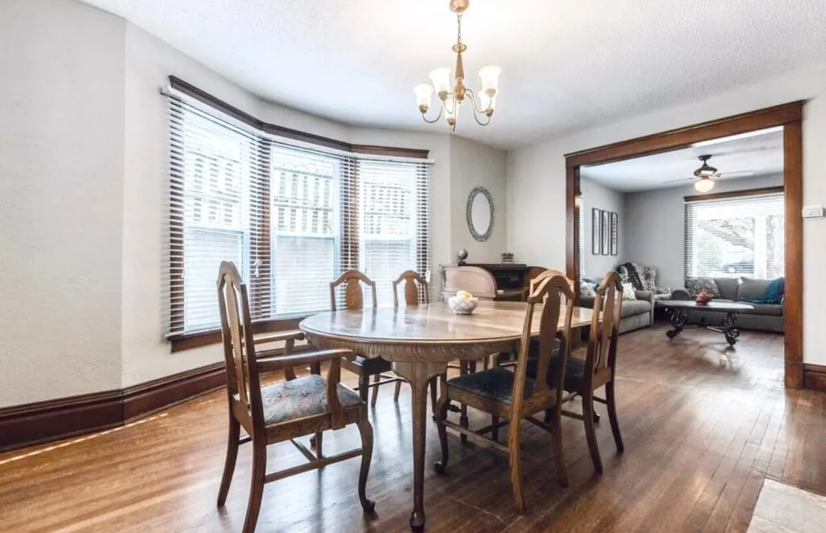 An inviting dining area featuring a large dining table beneath a sparkling chandelier, adjacent to a bay window that fills the space with natural light. The scene suggests a place for family meals, group gatherings, and warm conversations, capturing the essence of shared moments.