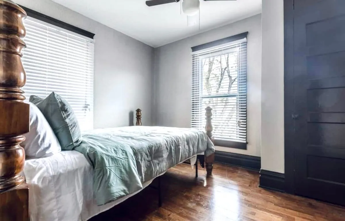 This serene bedroom features a bed dressed in light green bedding, a large window with blinds, and polished wooden flooring. The soft, natural lighting creates a calm and airy environment, perfect for recharging.