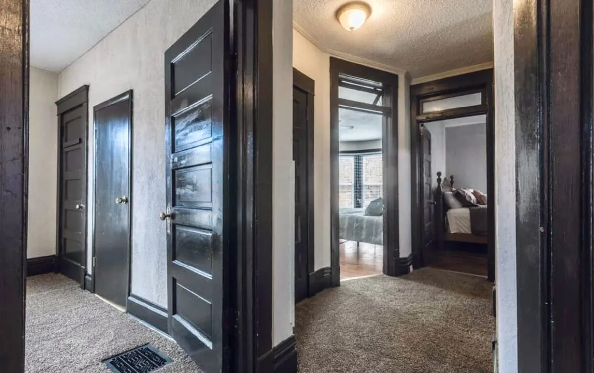 A transitional hallway space with dark wooden doors, carpeted flooring, and natural light spilling in from windows, creating a quiet, reflective atmosphere. It evokes movement and the idea of passing through, metaphorically connecting the home to the surrounding city.