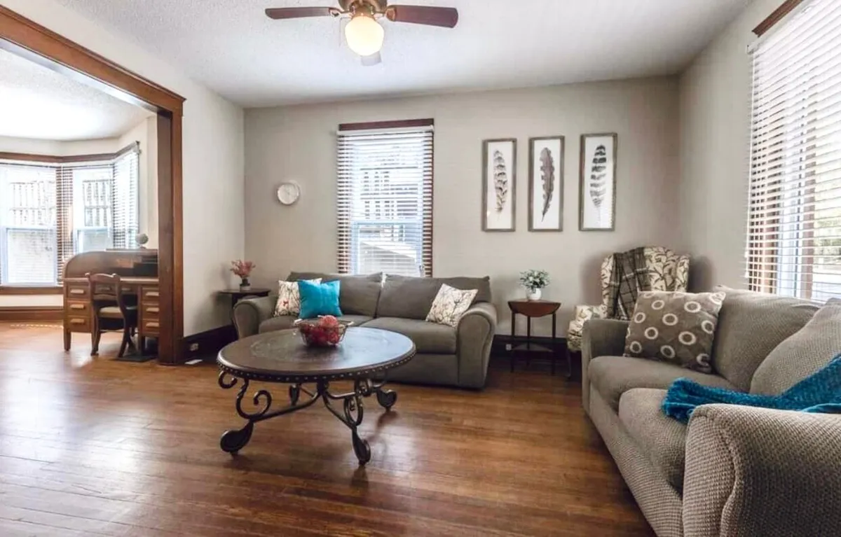 Another angle of the living area, featuring a coffee table, a comfortable sofa, artwork on the wall, and a window with blinds. The scene suggests both relaxation and social interaction, with space for reading, conversation, or casual gatherings.