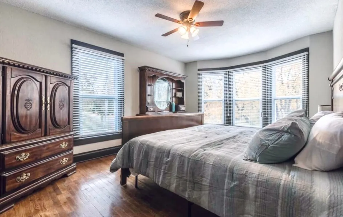 A tranquil bedrooms space with a wooden dresser, bay windows letting in plentiful daylight, and a neatly made bed with pillows. The room is filled with natural light, creating an ideal environment for quiet reflection, reading, or working in a serene setting.