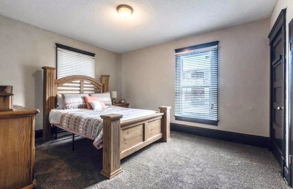 A cozy bedroom featuring a comfortable bed positioned by a bright window, complemented by a simple nightstand. The room is filled with natural light, creating an inviting and restful atmosphere—ideal for unwinding after a productive workday.