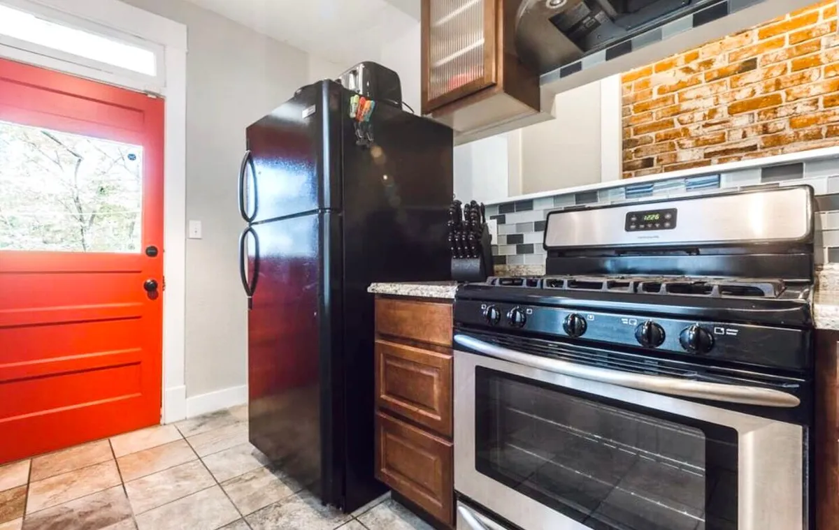 This image showcases a welcoming kitchen with a red front door, black refrigerator, stainless steel oven, granite countertop, and a brick backsplash. The scene is lively, suggesting the space is ready for cooking, entertaining, and family gatherings. All major kitchen appliances and surfaces are clearly visible, emphasizing functionality and warmth.